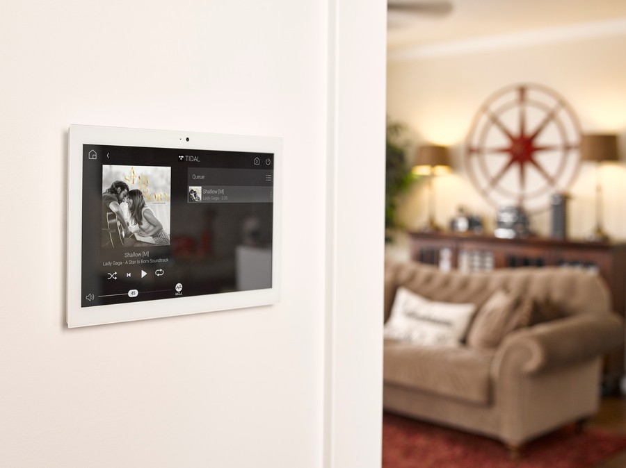 A Control4 wall panel in a living room displaying the song "Shallow" by Lady Gaga